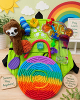 A bright green sensory backpack filled with colorful play accessories, including a plush sloth, a rainbow maze, a blue striped sensory stick, a yellow star, a clear marble run, and a lava tube, arranged on a light wood surface with decorative background elements.